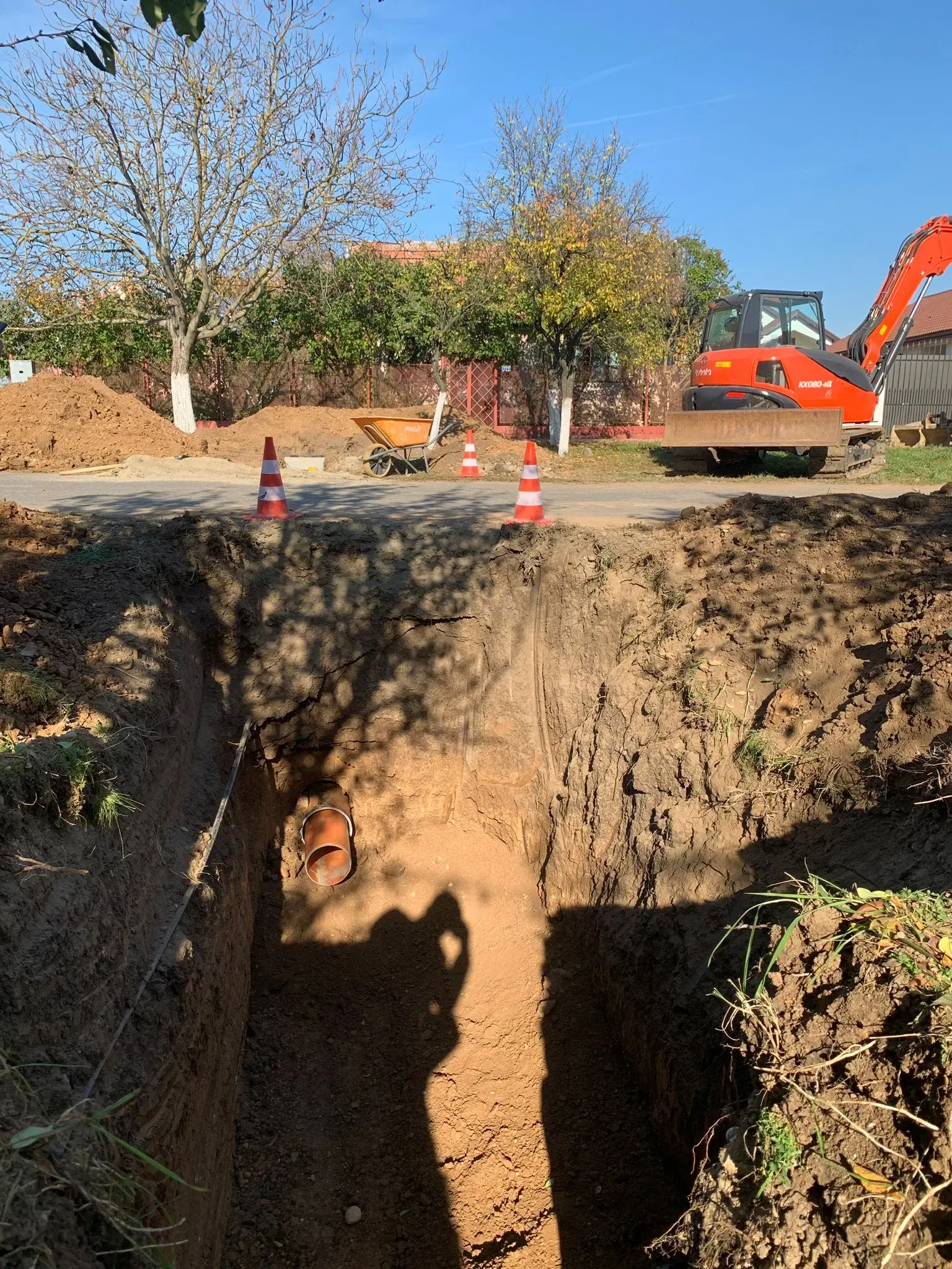 Șantier de branșament la marginea drumului, cu excavatorul poziționat lângă tranșee