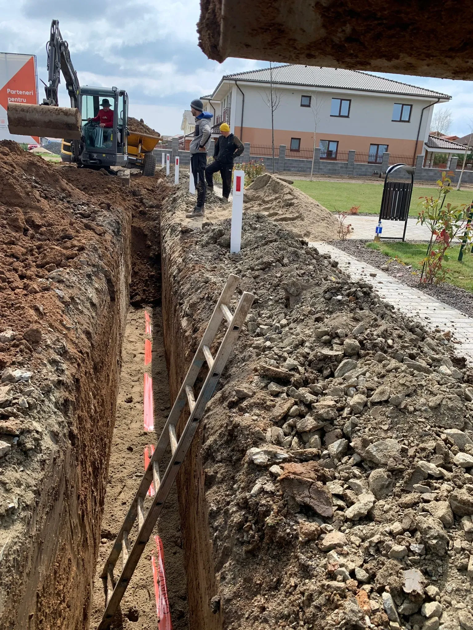 Săpătură pentru racord în fața unei case noi, cu miniexcavator și scară de acces în tranșee